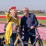 Een lachend ouder echtpaar staat met fietsen terwijl ze een selfie maken in een levendig tulpenveld. De man draagt een blauwe jas en een bril, en de vrouw draagt een gele jas met een sjaal en houdt een rode telefoon vast. Het veld staat vol met rijen roze en rode tulpen.