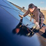 Een arbeider in een harnas installeert zonnepanelen op een dak van een betegeld huis. Hij gebruikt een boormachine, draagt een pet en handschoenen, met een helderblauwe lucht op de achtergrond.