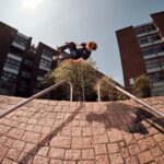 Een skateboarder voert een truc uit boven een leuning op een met bakstenen geplaveide helling in een stedelijke omgeving. Twee mensen staan erbij en kijken ernaar, met moderne gebouwen en een boom op de achtergrond onder een heldere hemel.