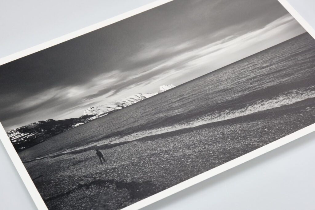 Een zwart-witfotoafdruk toont een eenzame figuur die op een kiezelstrand staat. De oceaan strekt zich uit naar de horizon onder een bewolkte hemel. In de verte zijn met sneeuw bedekte bergen zichtbaar, die de zee ontmoeten. De foto wordt op een witte ondergrond geplaatst.