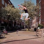 Een persoon is in de lucht, springt van een richel op een straat, met opgeheven armen en gebogen benen. Een ander persoon staat op de richel, houdt een camera of telefoon vast en legt de actie vast. Fietsen staan in de buurt geparkeerd en bomen met witte bloesems omzomen de woonstraat.